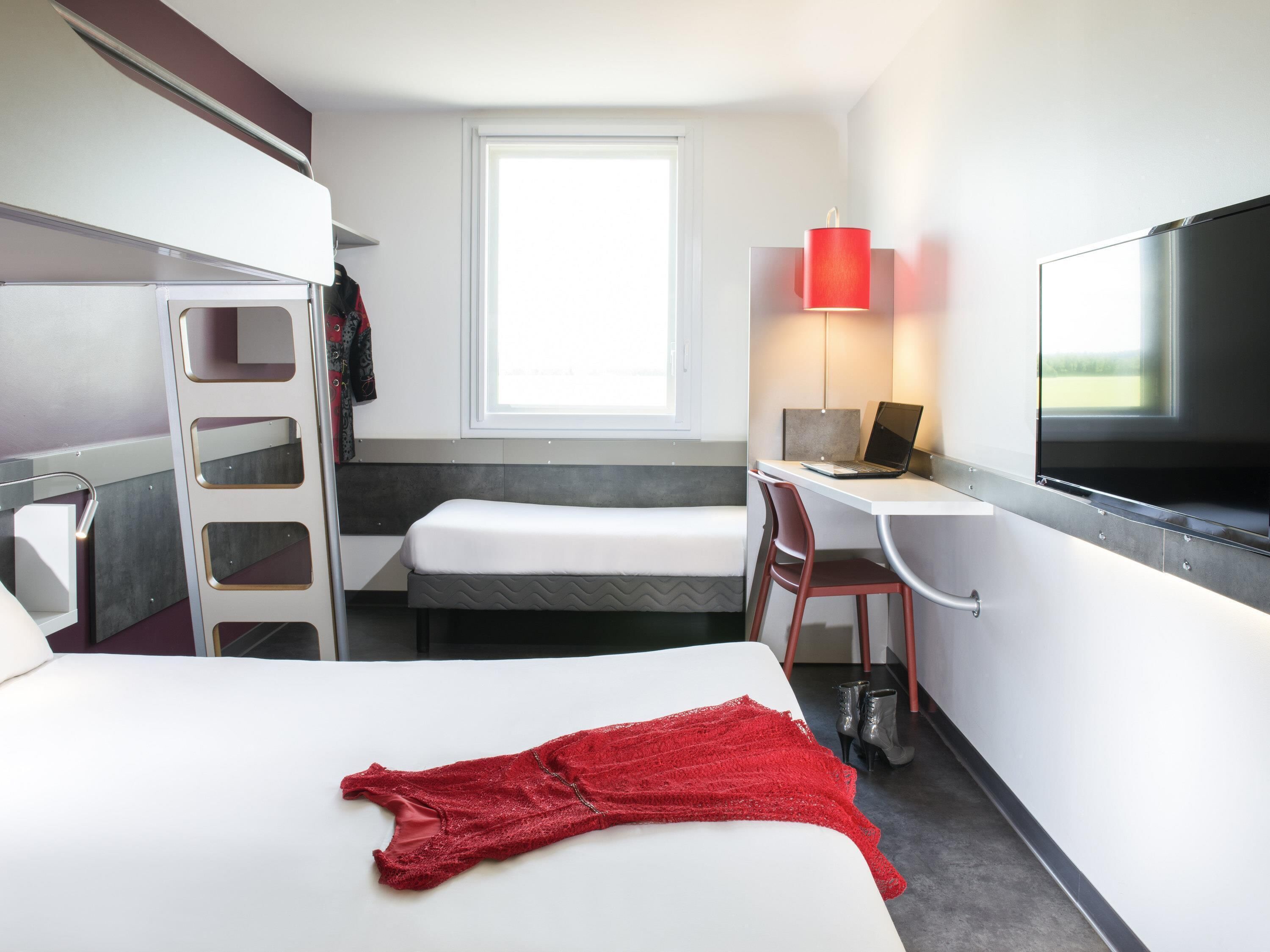 A hotel room with a bed, a desk with a laptop, a TV, a window, and a red garment on the bed.