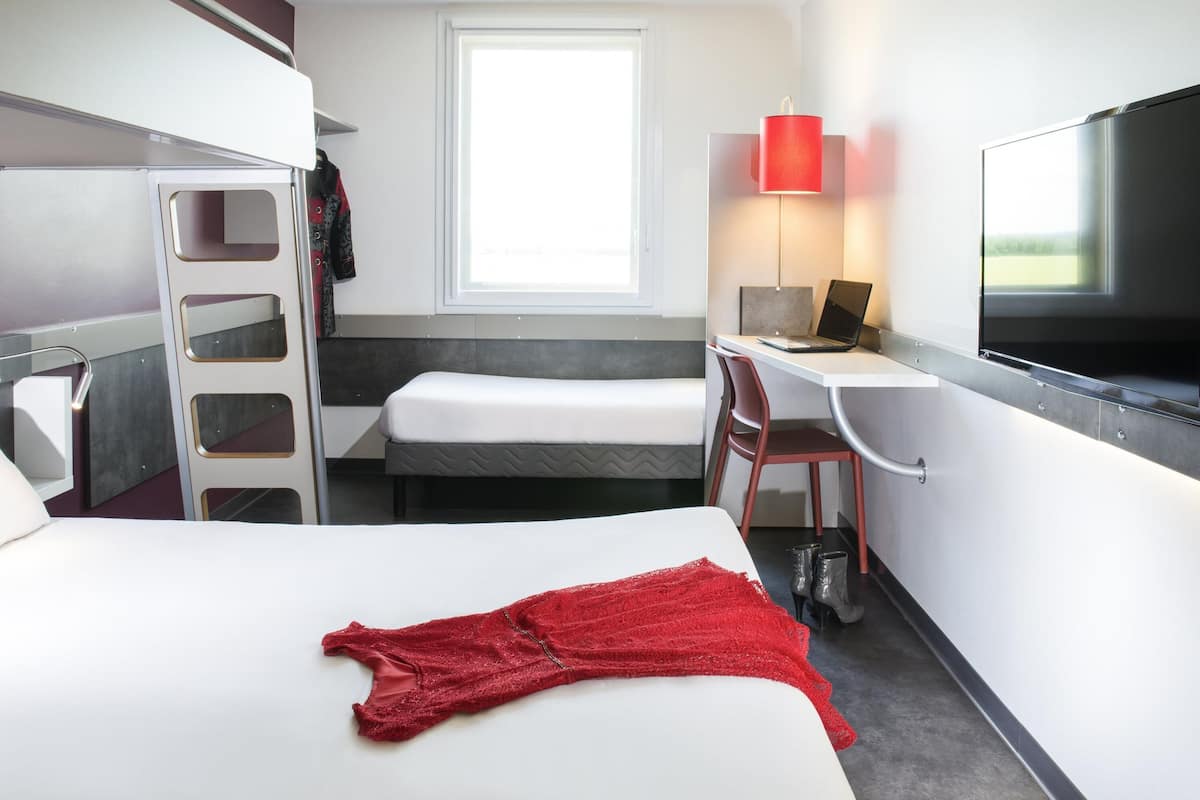 Une chambre d’hôtel avec un lit, un bureau avec un ordinateur portable, une télévision, une fenêtre et un vêtement rouge sur le lit.