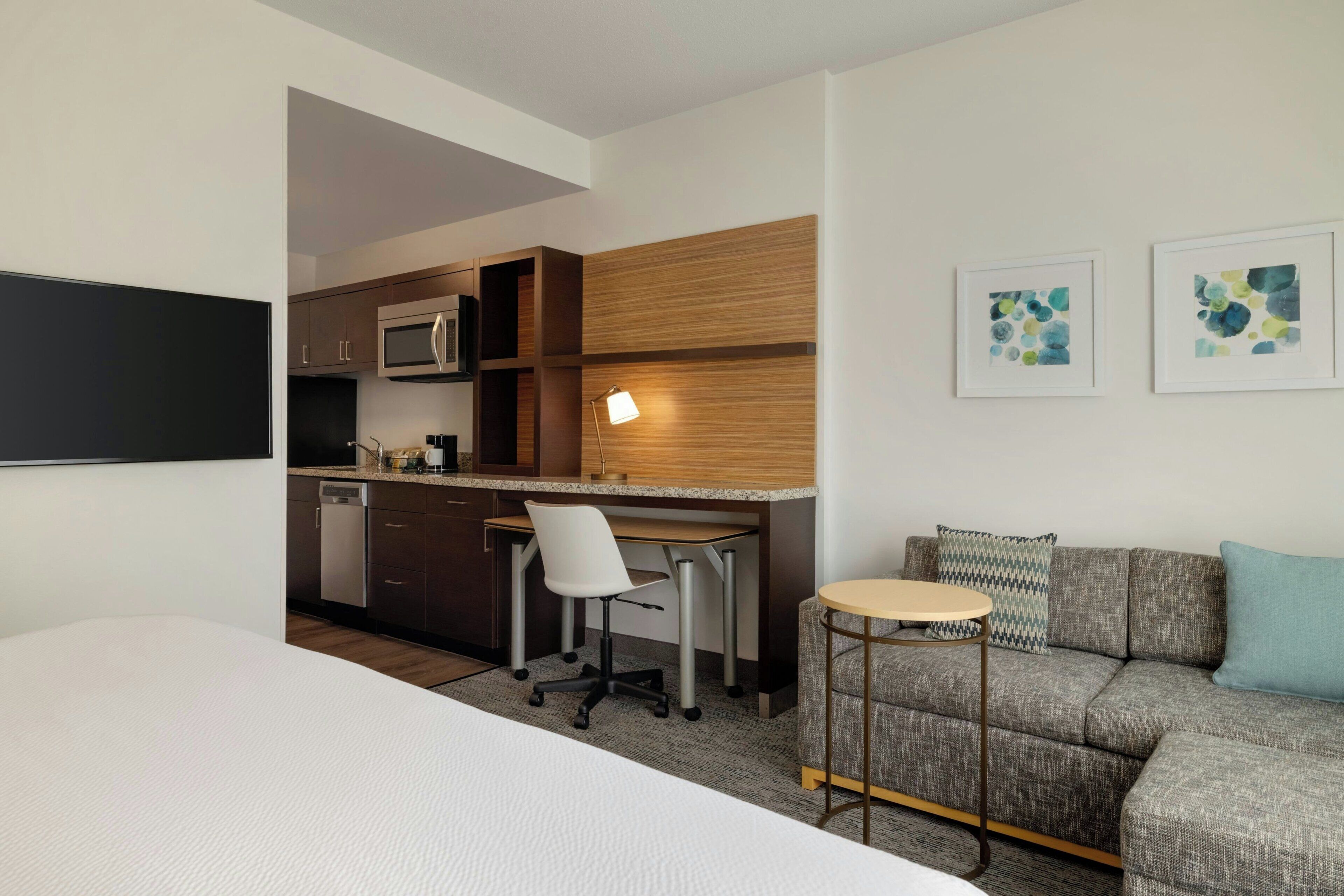 Een compacte hotelkamer met kitchenette, een bank, een klein rond tafeltje en twee ingelijste kunstwerken aan de muur.
