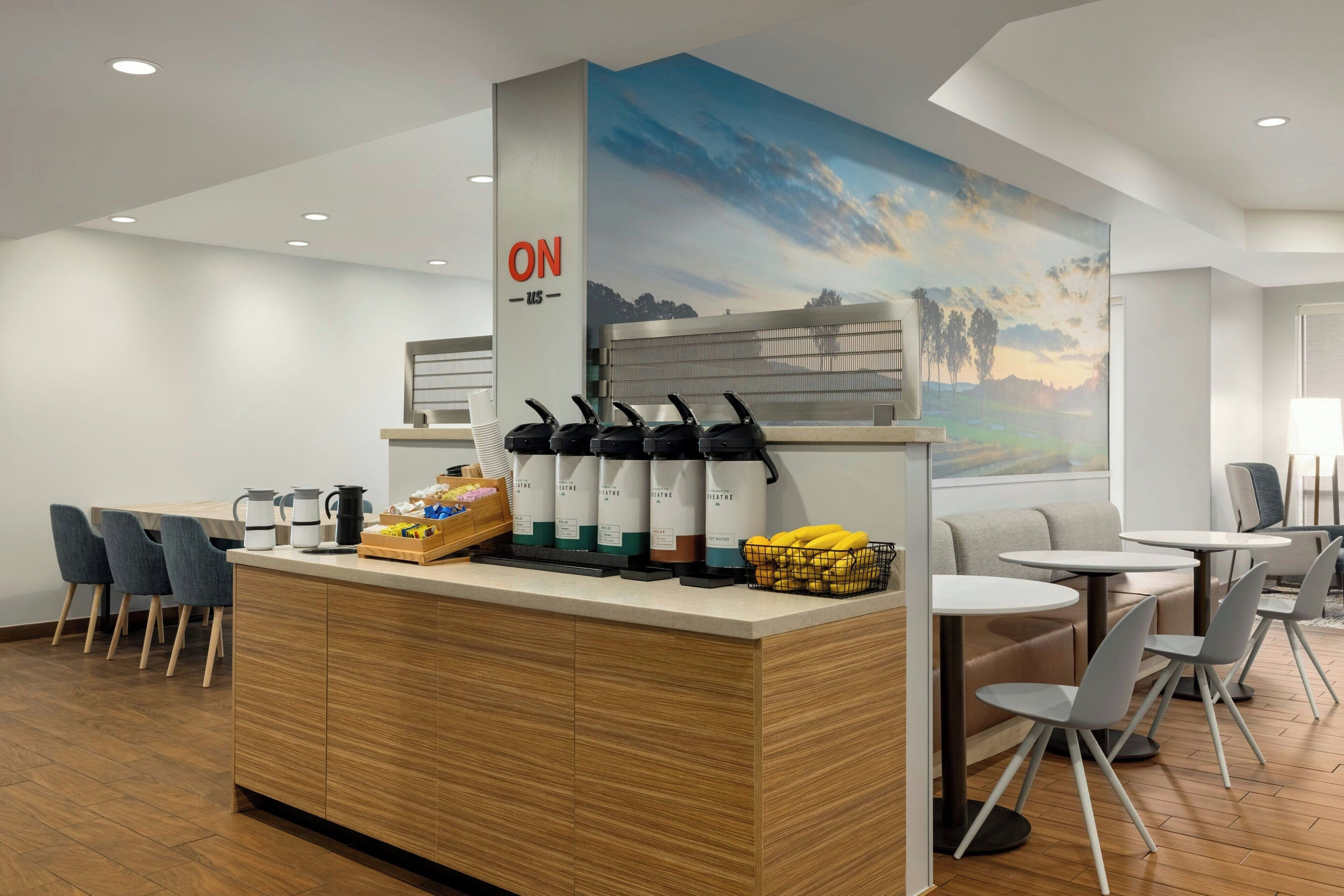 A modern coffee station with a wooden counter, a painting of a landscape, and seating areas with tables and chairs.