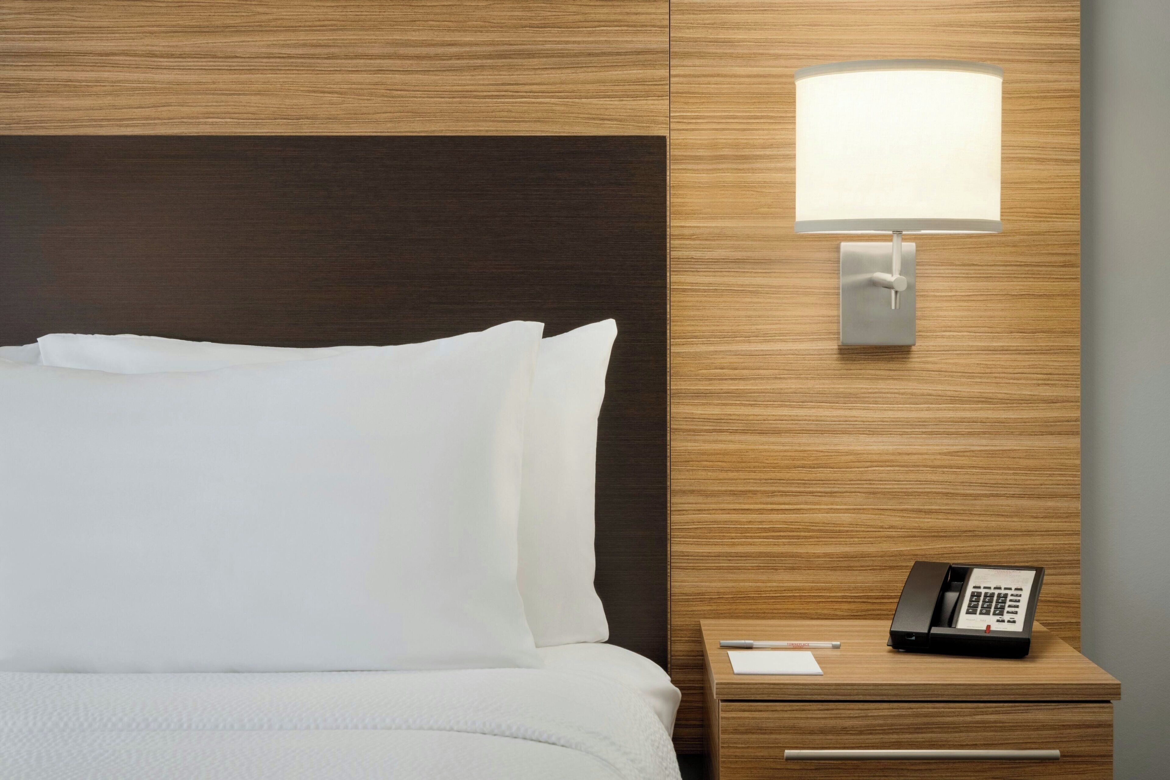 Una habitación de hotel con una cama bien hecha, una lámpara de noche y un teléfono sobre una mesita de madera.