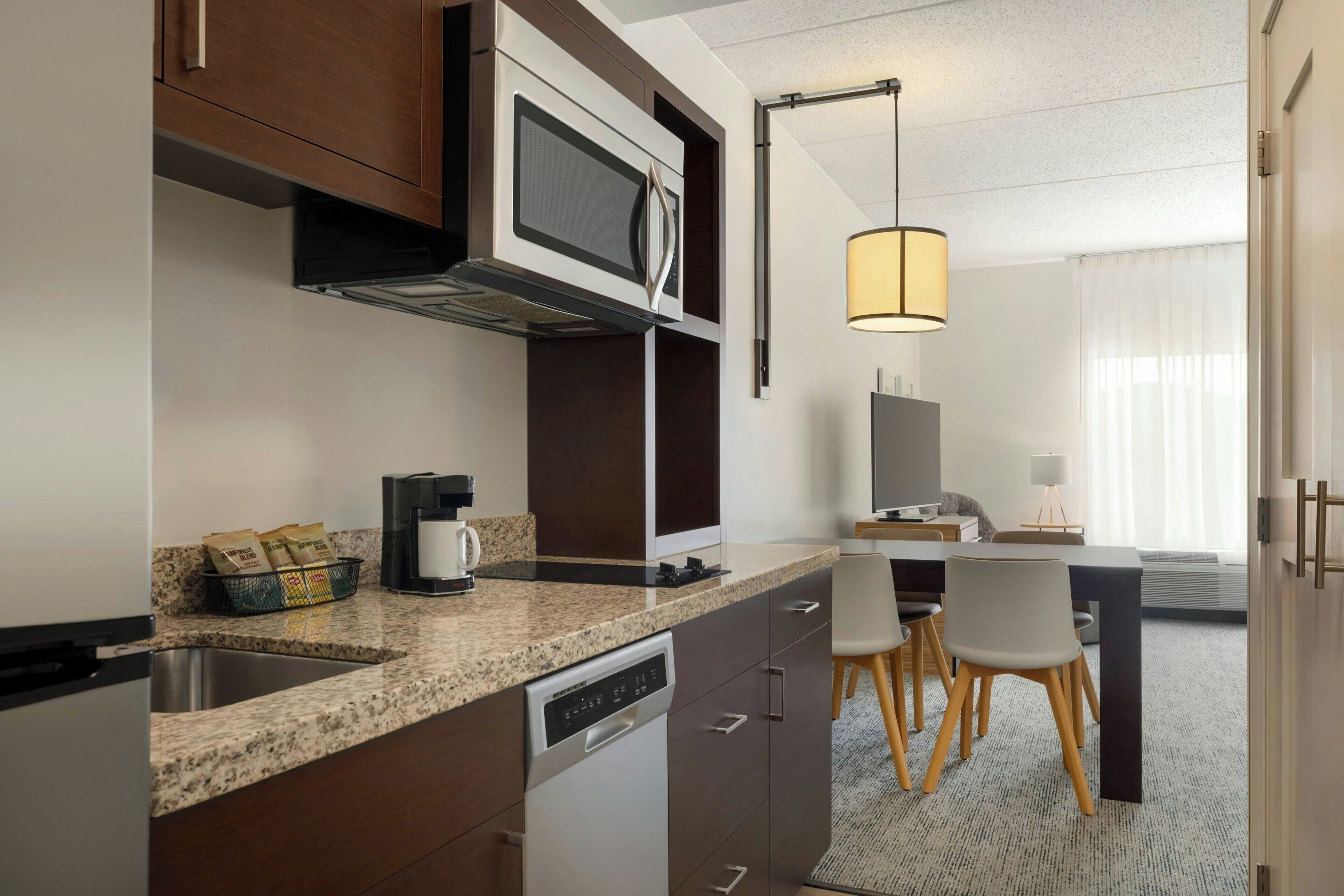 Una cocina de hotel moderna con encimeras de granito, microondas integrado, cafetera y lavavajillas. Hay un comedor con mesa y sillas, y un amplio ventanal con cortinas.