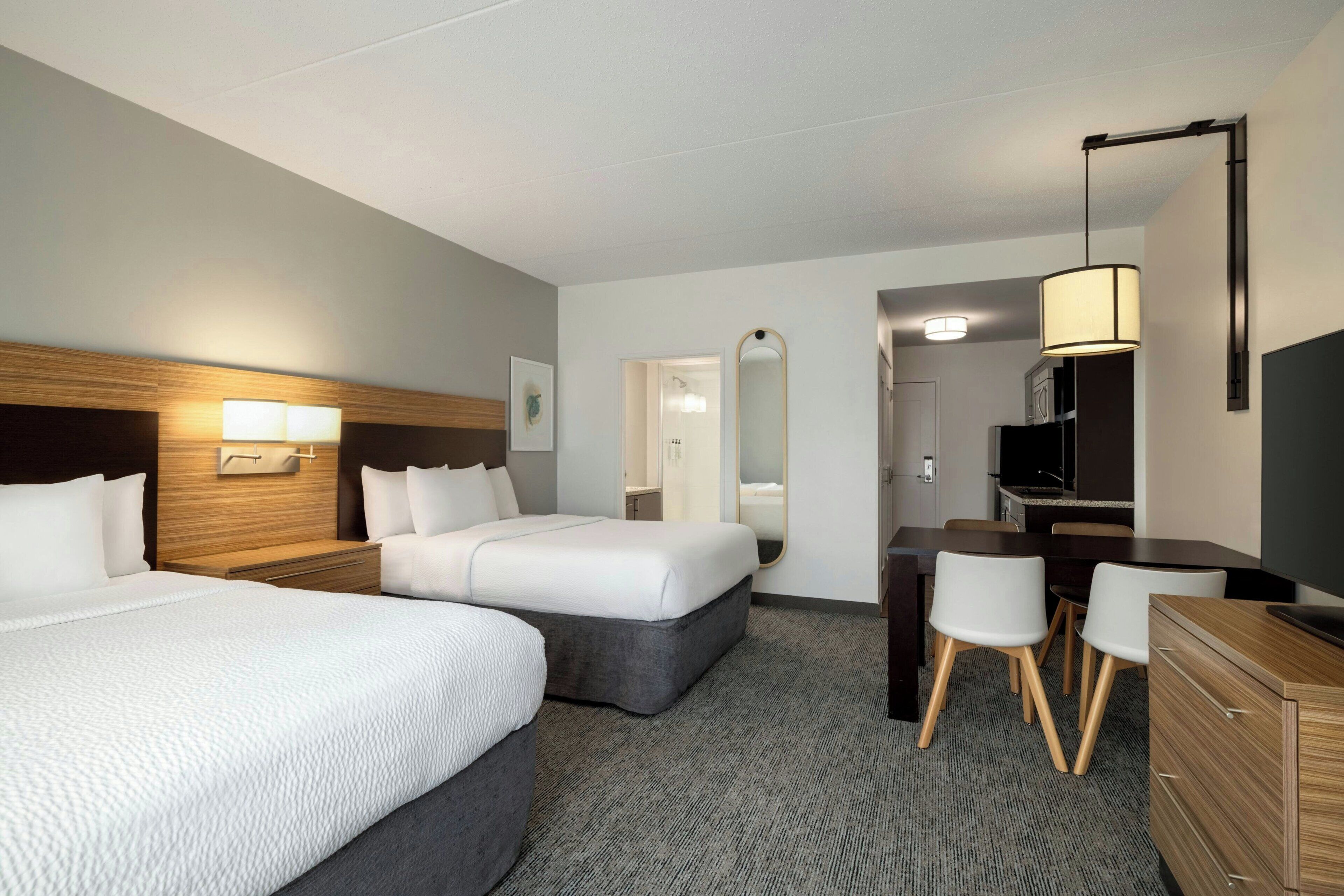 A hotel room with two beds, a desk, chairs, a television, and a mirror.