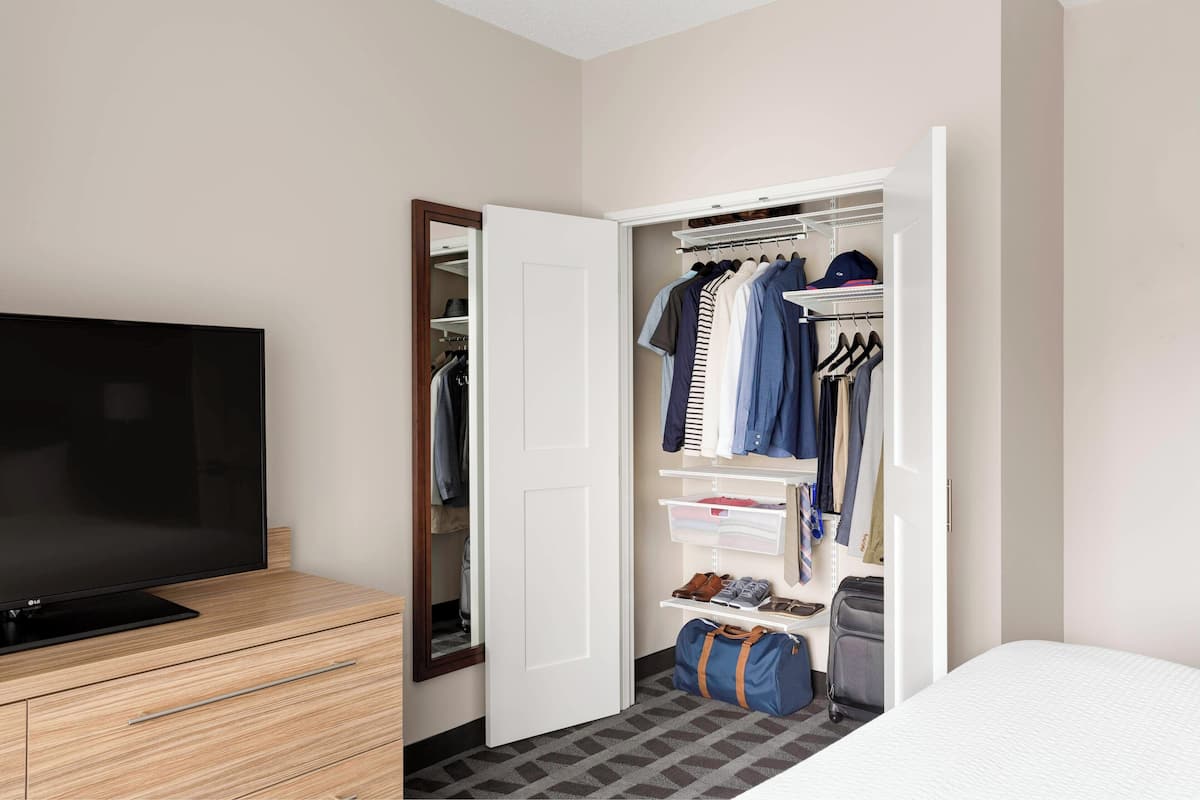 A hotel room with a TV on a wooden dresser, a walk-in closet with hanging clothes and shelves, and a bed with a white comforter.