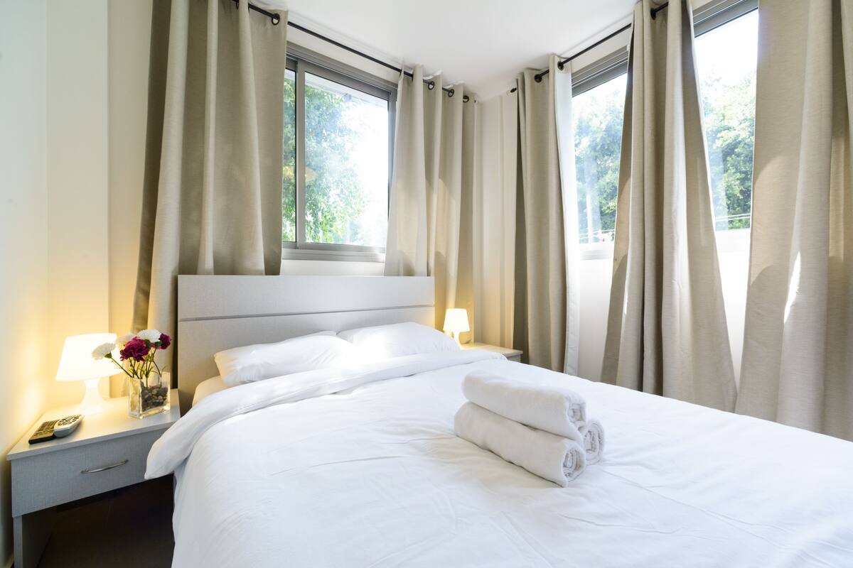 Ein Hotelzimmer mit einem ordentlich bezogenen Bett, einem Nachttisch mit Lampe und Blumenvase sowie großen Fenstern mit Vorhängen.