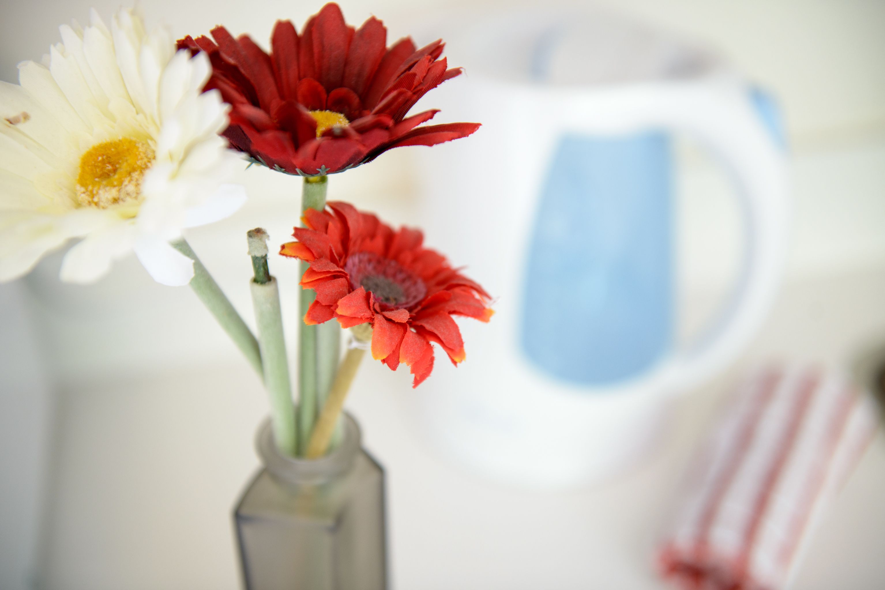 A vase with three flowers and a white pitcher in the background.