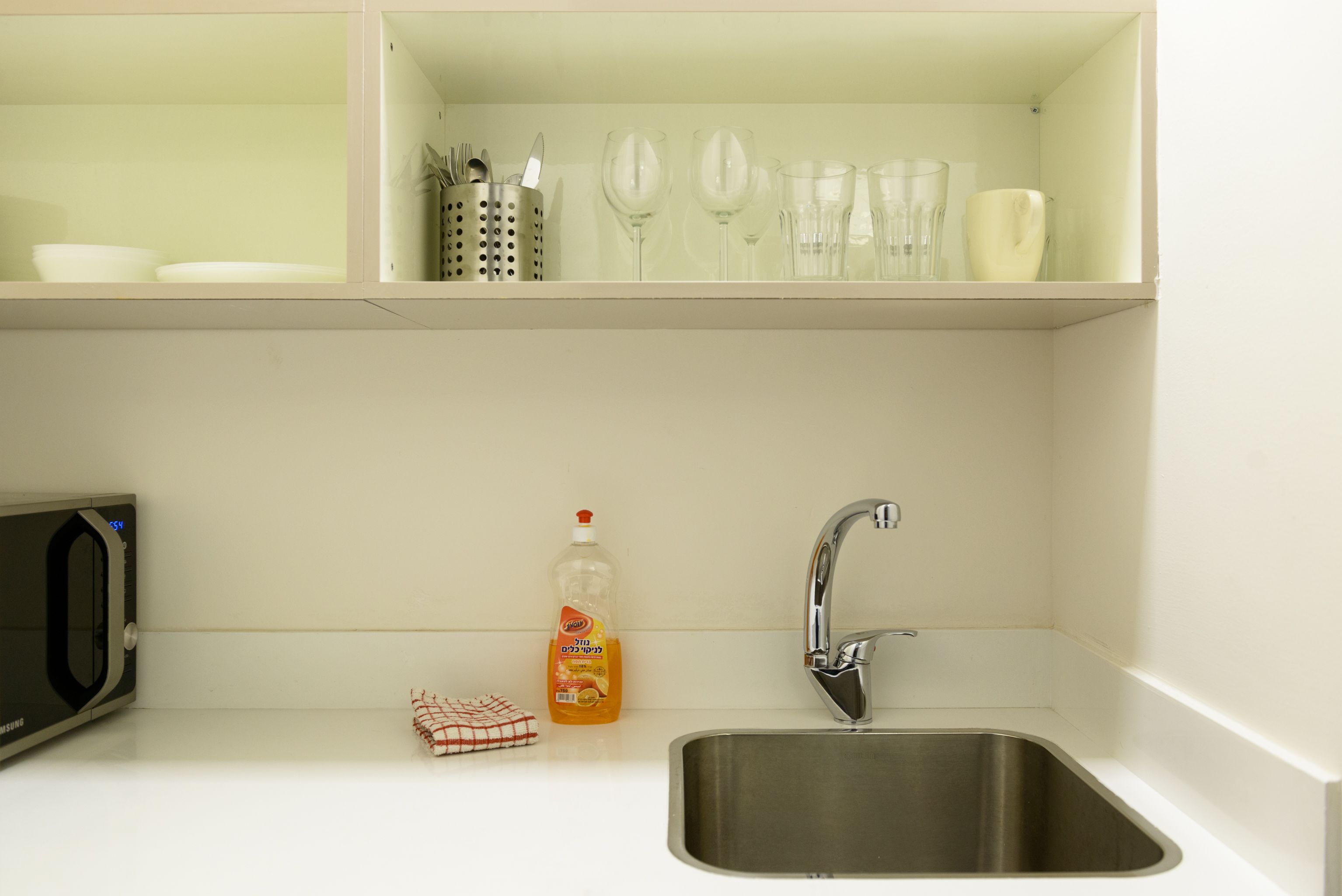 A kitchen with a stainless steel sink, a microwave, and a shelf above the sink holding glassware and utensils.
