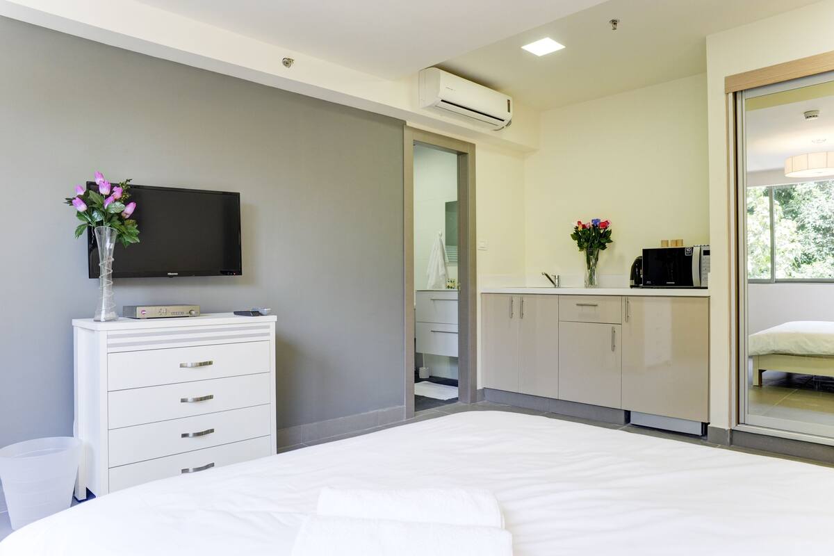 Ein modernes Hotelzimmer mit Bett, Fernseher auf einer Kommode, einem durchgegangenen Tür erkennbaren Badezimmer und einem Fenster mit Blick auf Grünflächen.