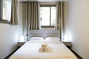 Una cama bien hecha con toallas enrolladas, una mesita de noche con una lámpara y cortinas en una ventana.