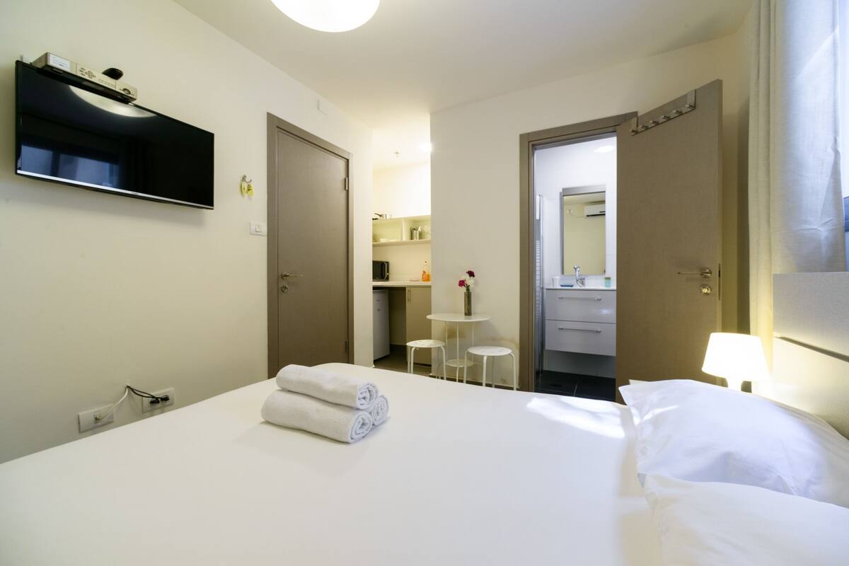 Ein Hotelzimmer mit Bett, einem an der Wand befestigten Fernseher, einem Schreibtisch mit Stuhl und einem durchgehenden Türspalt einsehbarer Badezimmer.