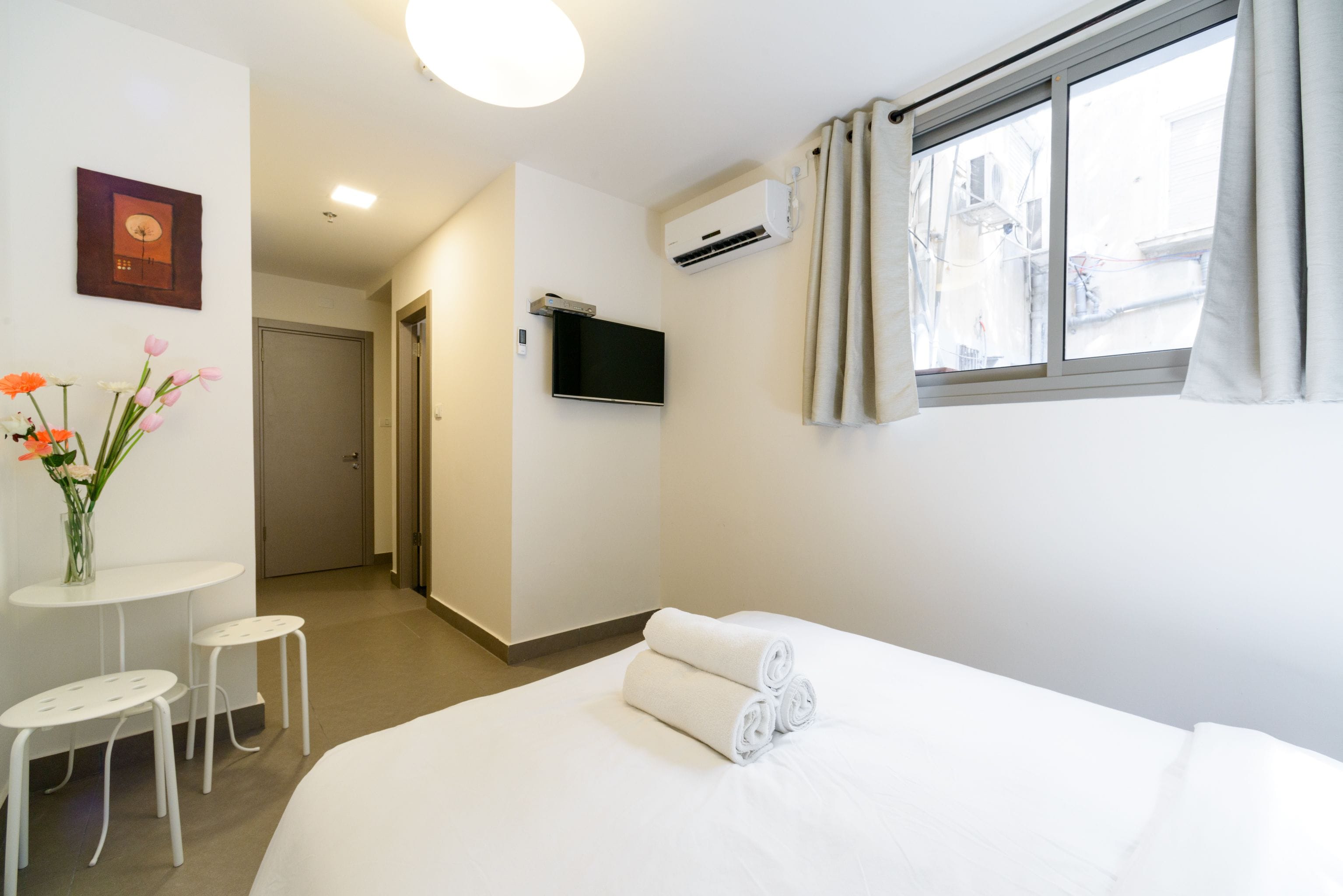 A hotel room with a bed, a TV, an air conditioning unit, a window with curtains, a small table with two chairs, and a vase with flowers.