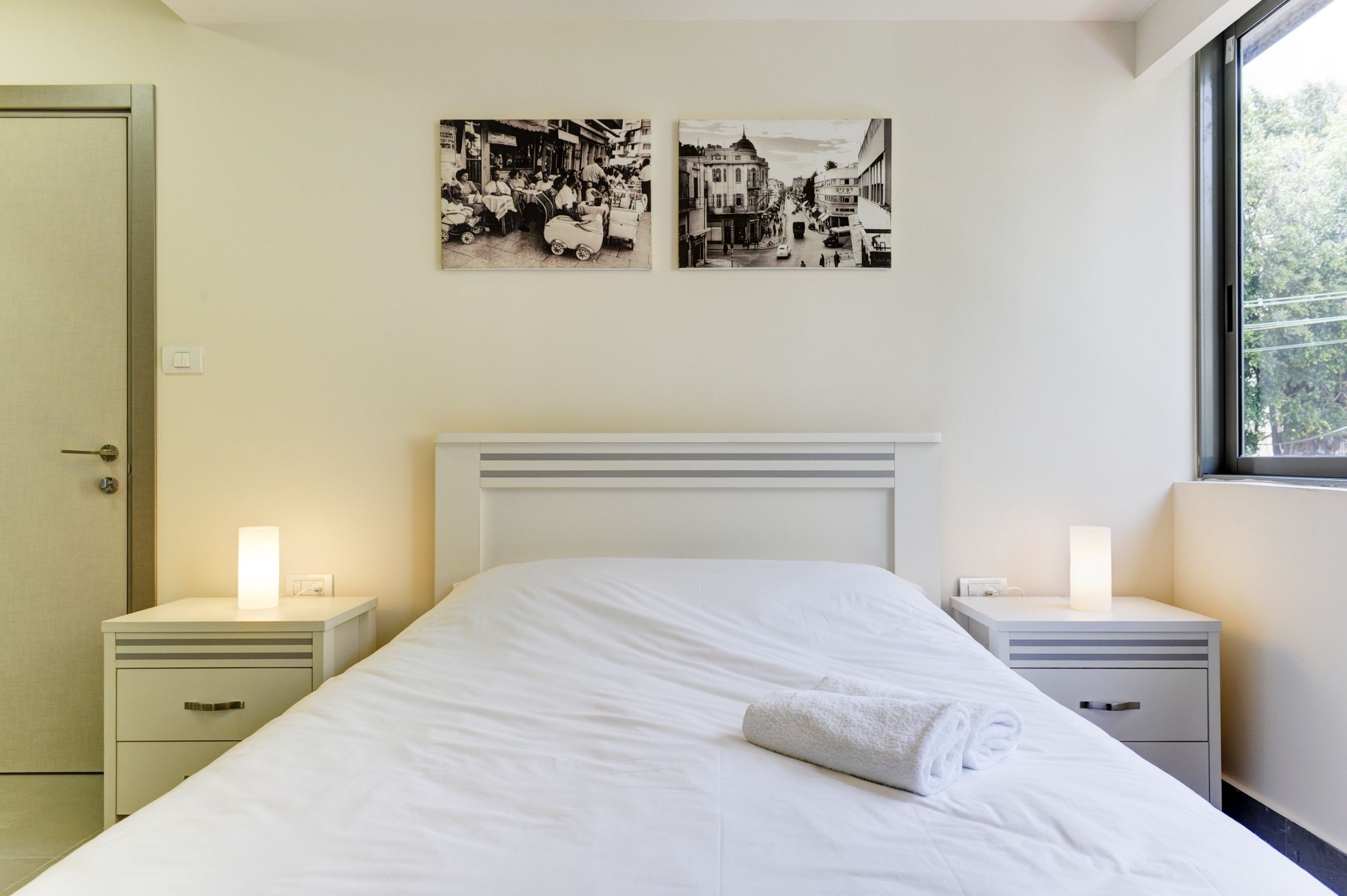 Ein Hotelzimmer mit einem großen Bett, zwei Nachttischen mit Lampen, einer Tür und zwei schwarz-weißen Fotografien an der Wand.