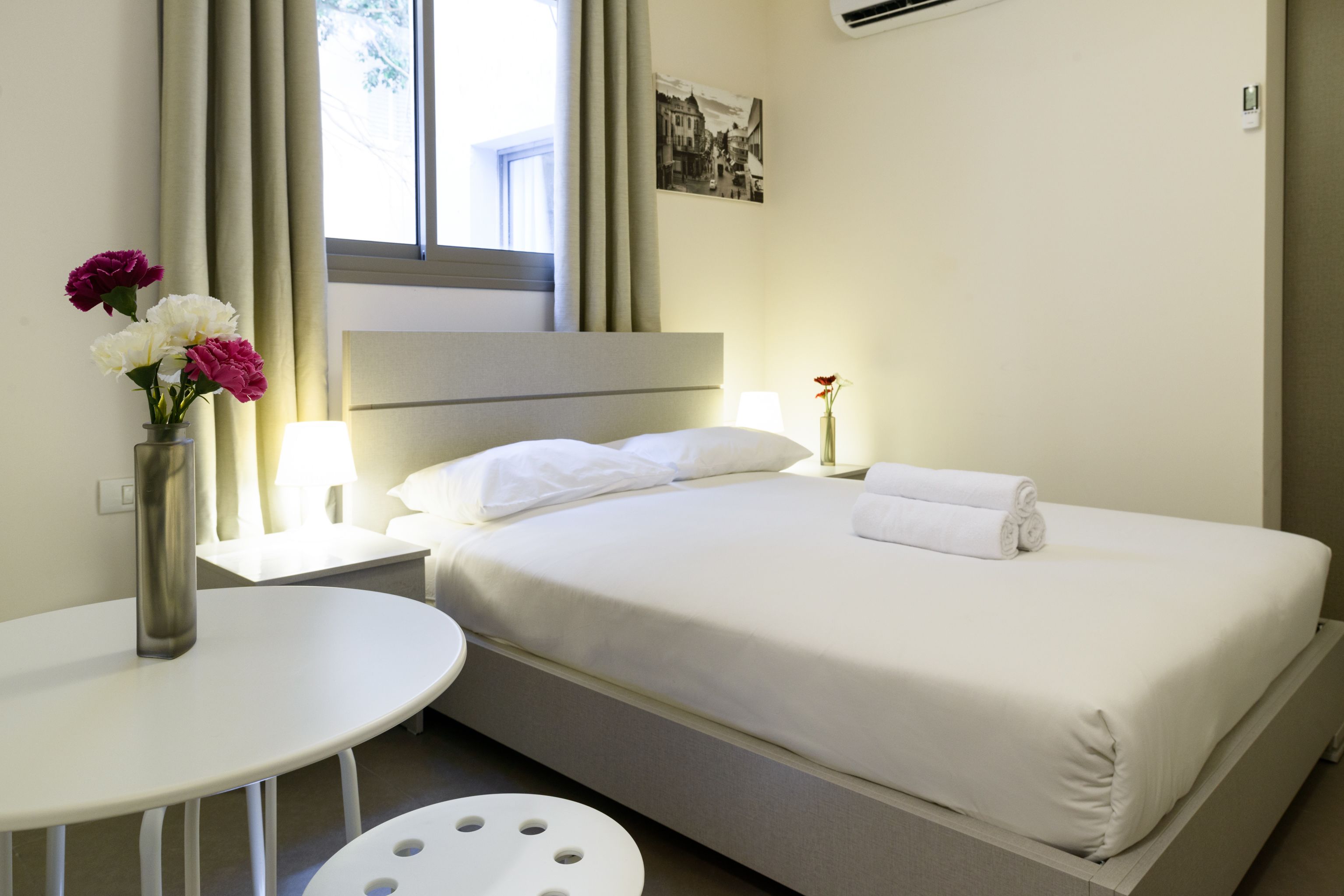A hotel room with a bed, a bedside table with a lamp, a small round table, and a vase with flowers.