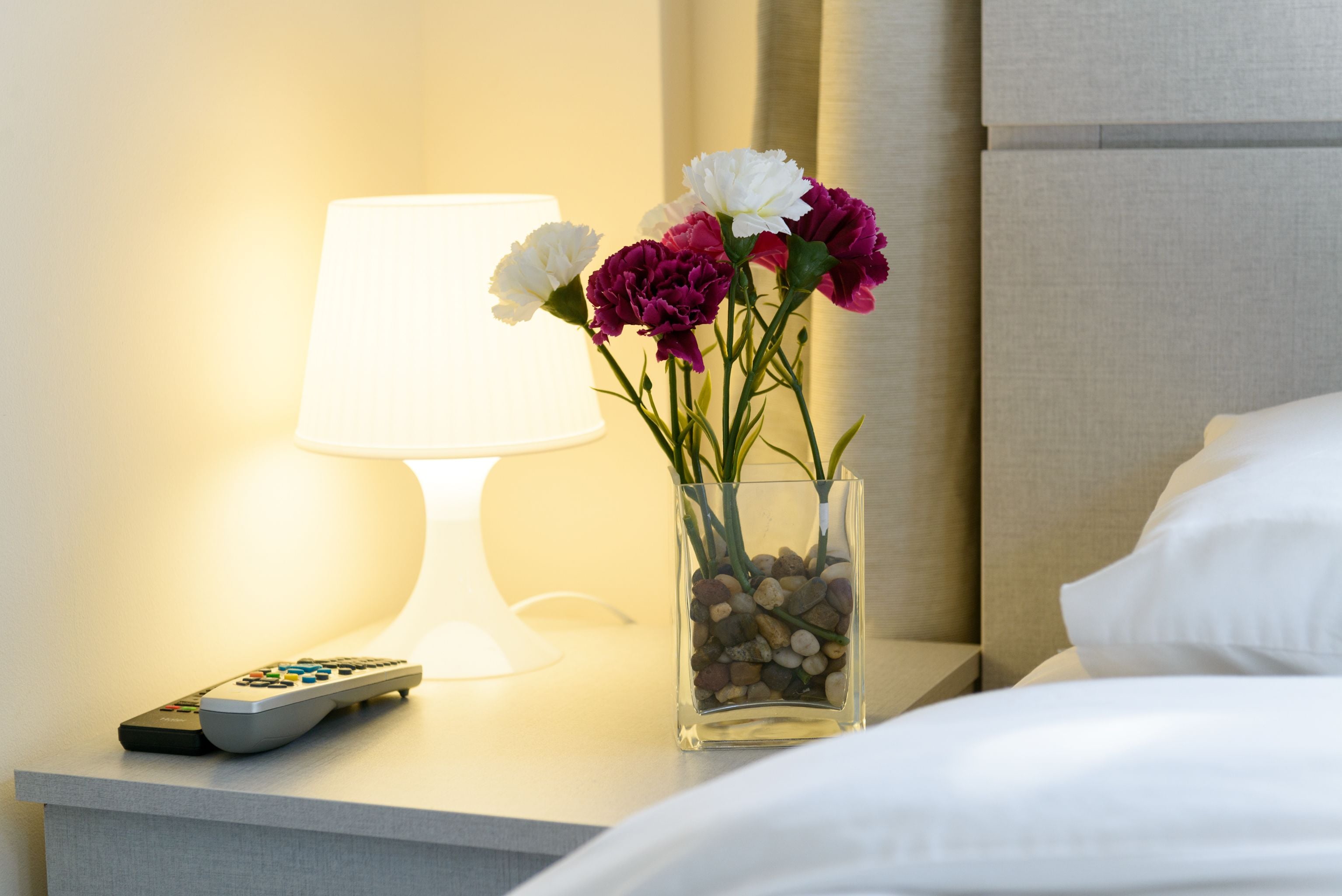 A bedside table with a remote control, a lamp, and a vase with flowers.