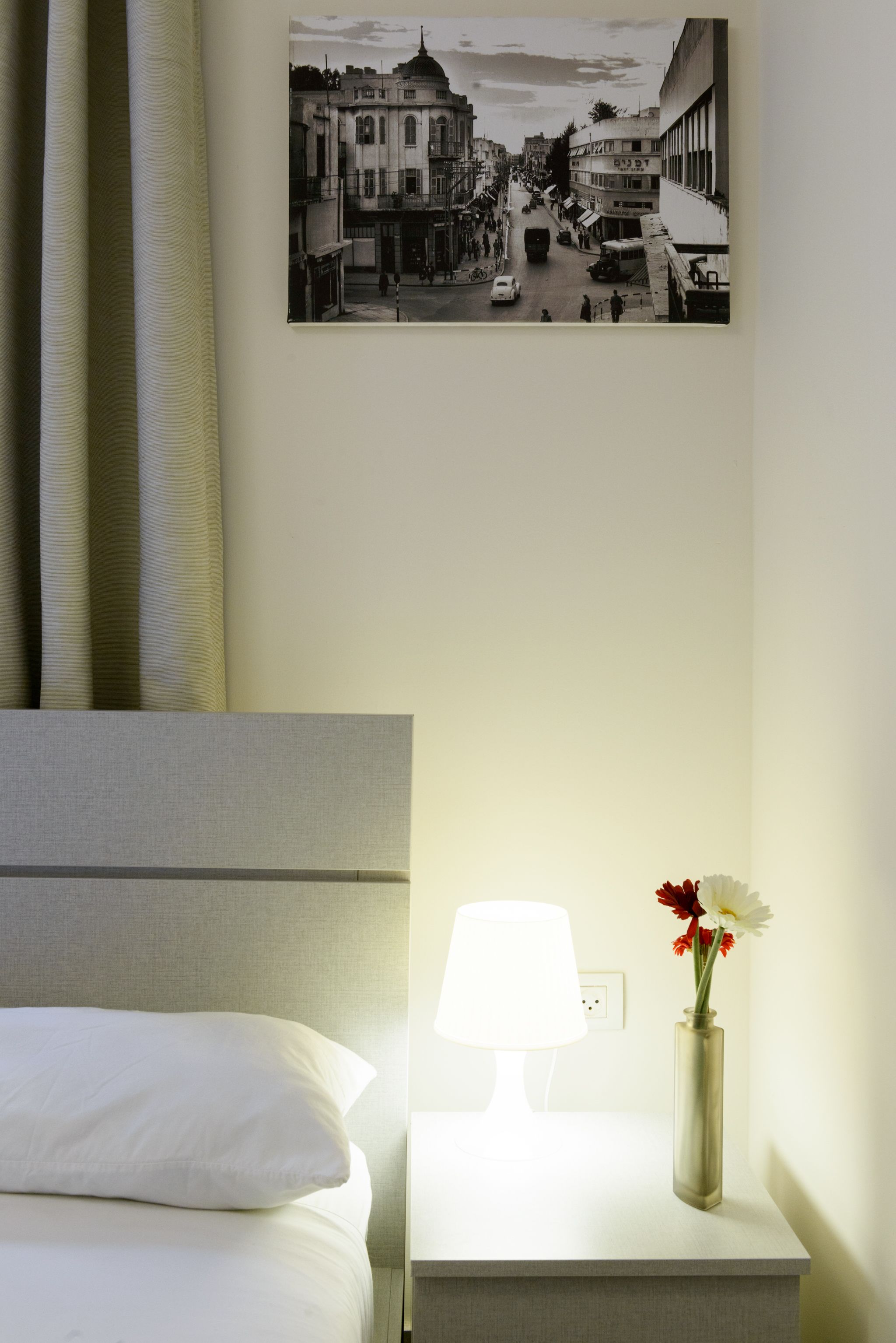 A hotel room with a bed, a nightstand with a lamp, and a wall-mounted picture of a street scene.