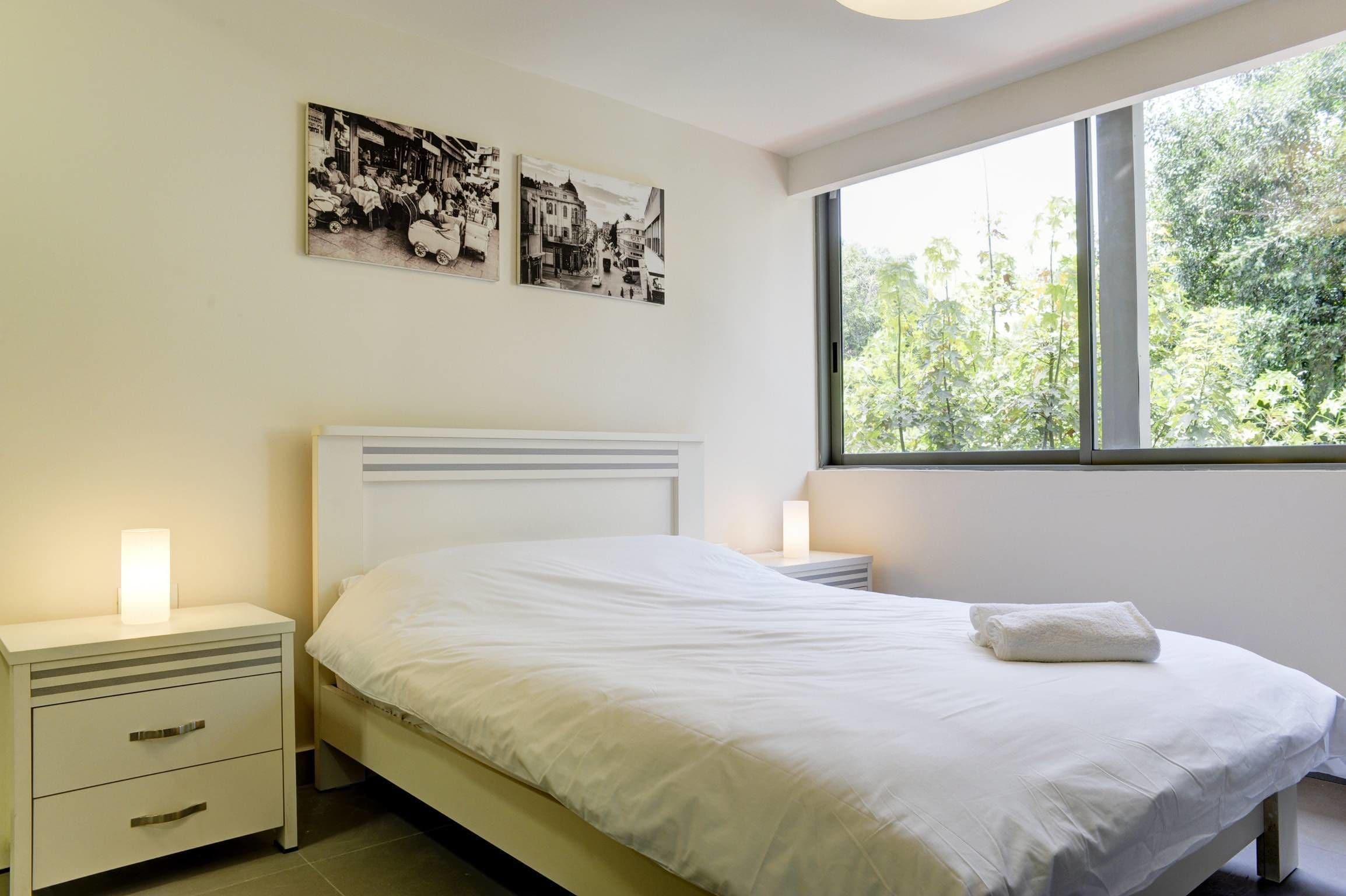 A bedroom with a bed, bedside lamps, a nightstand, and a window with a view of trees.