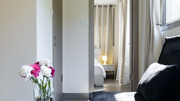 A hotel room with a bed, a bedside table with a vase of flowers, and a view into the adjacent room. - Dizengoff Inn (Tel Aviv)