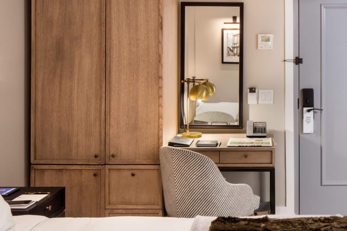 A hotel room with a wooden wardrobe, a desk with a lamp, a chair, and a bed.
