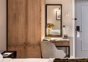 A hotel room with a wooden wardrobe, a desk with a lamp, a chair, and a bed.
