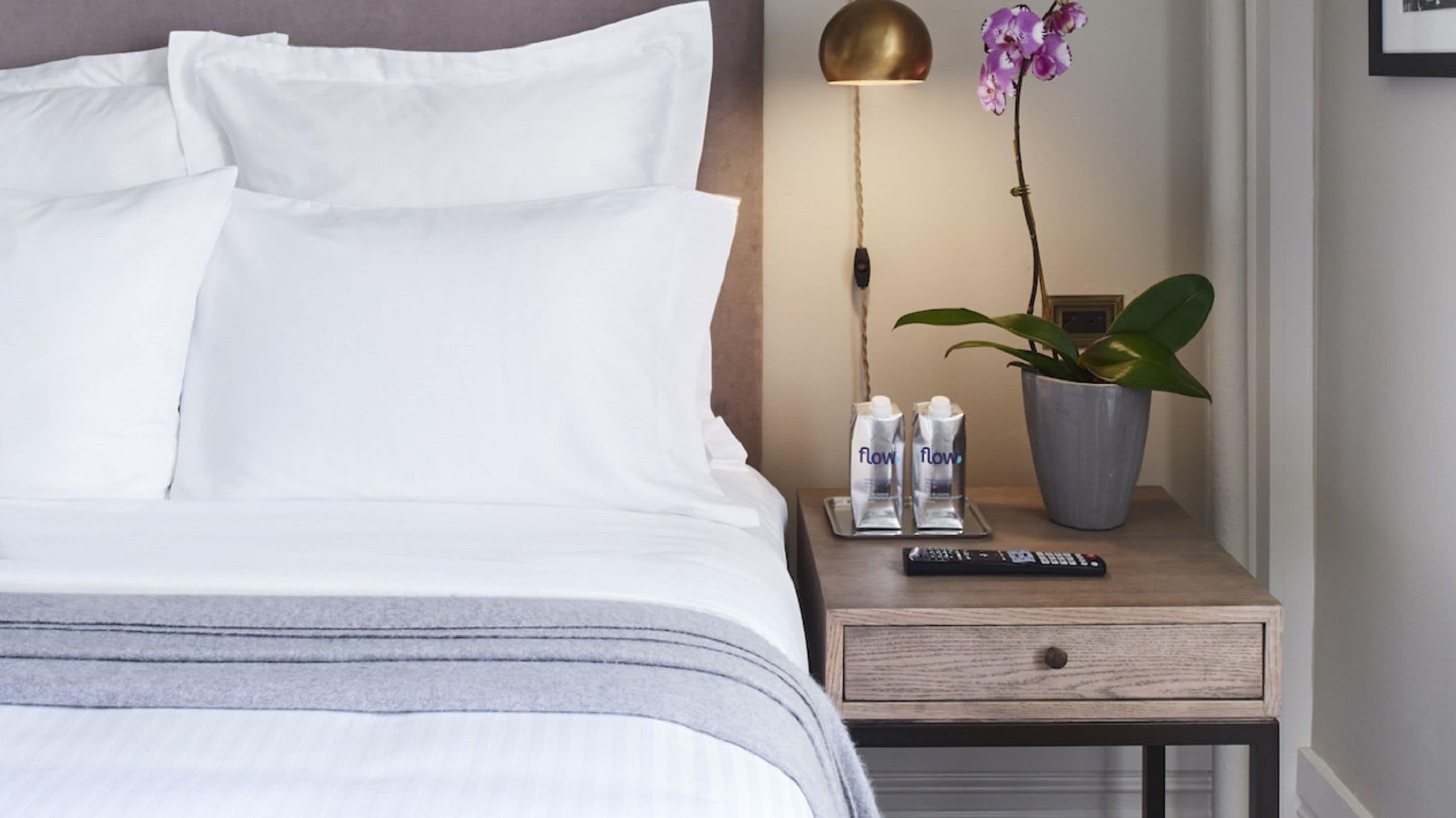 A neatly made bed with white linens, a bedside table with a remote control, a potted orchid, and two bottles labeled 'flow'.
