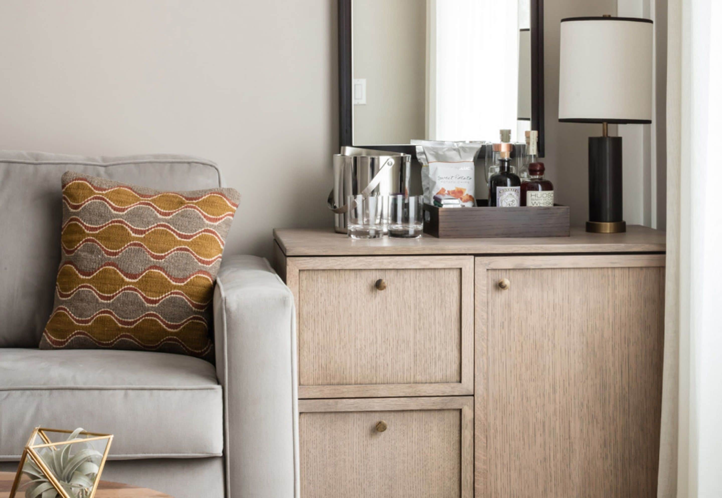 Habitación de hotel con un sofá gris, un cojín estampado, una mesita de madera con lámpara, un espejo y un minibar con distintos artículos.