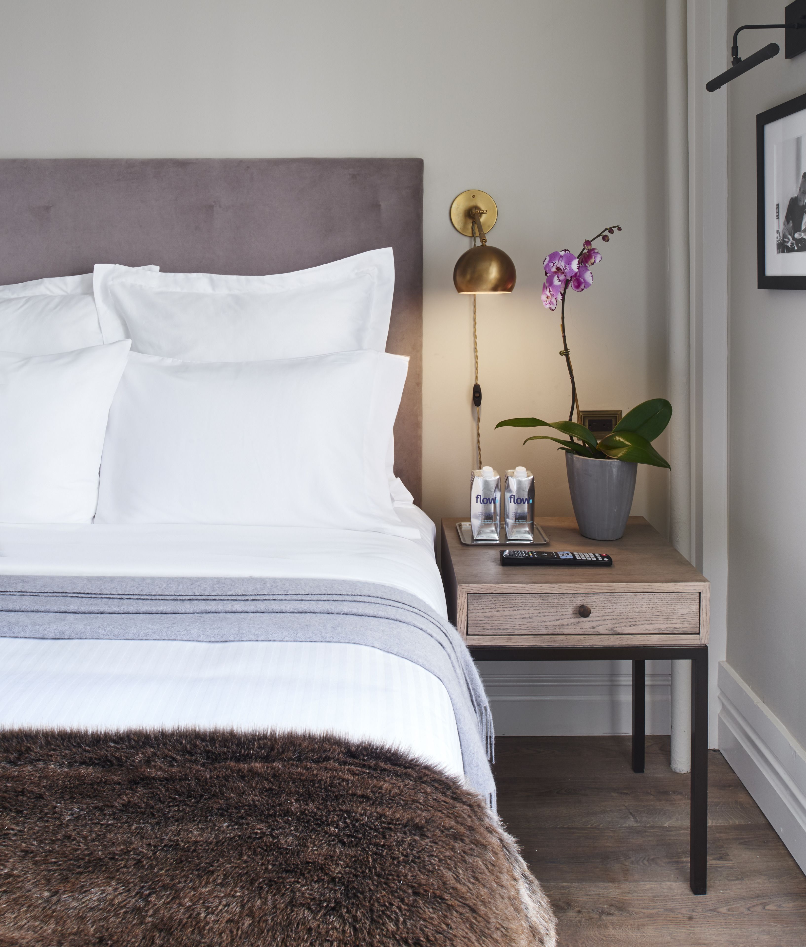 Una cama bien hecha con ropa blanca, una mesita de noche con una lámpara, una planta en maceta y un control remoto.