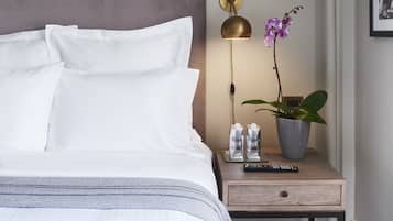Una cama bien hecha con ropa blanca, una mesita de noche con una lámpara, una planta en maceta y un control remoto.
