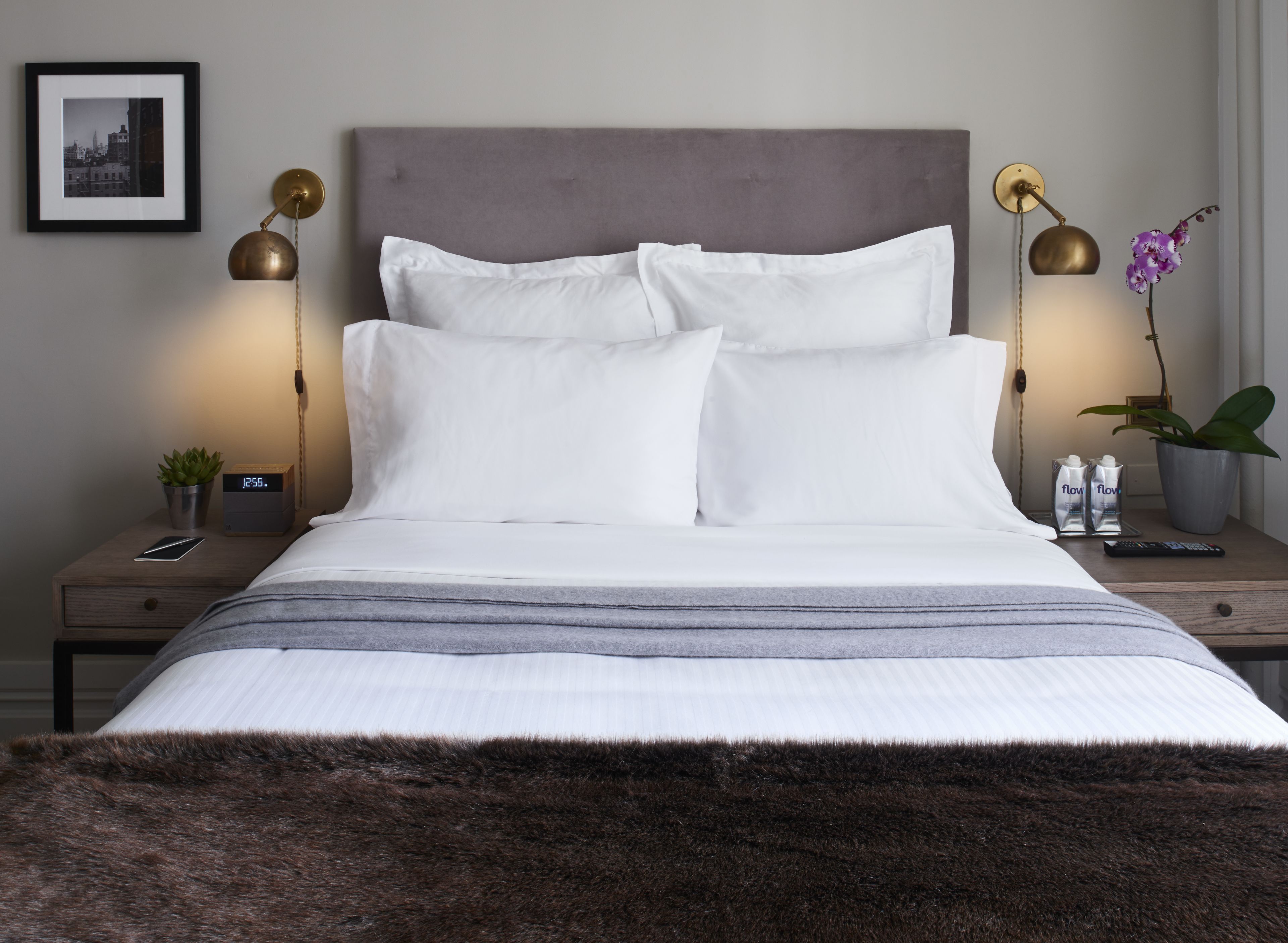 A neatly made bed with white linens and a grey headboard, flanked by two bedside tables with lamps and a small potted plant.