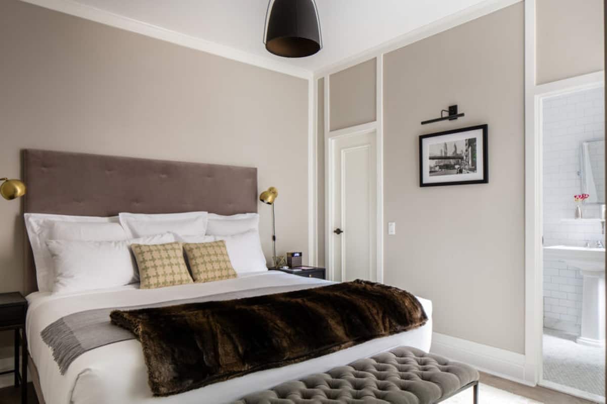 A bedroom with a large bed, a headboard, bedside lamps, a framed picture on the wall, and a door leading to a bathroom.