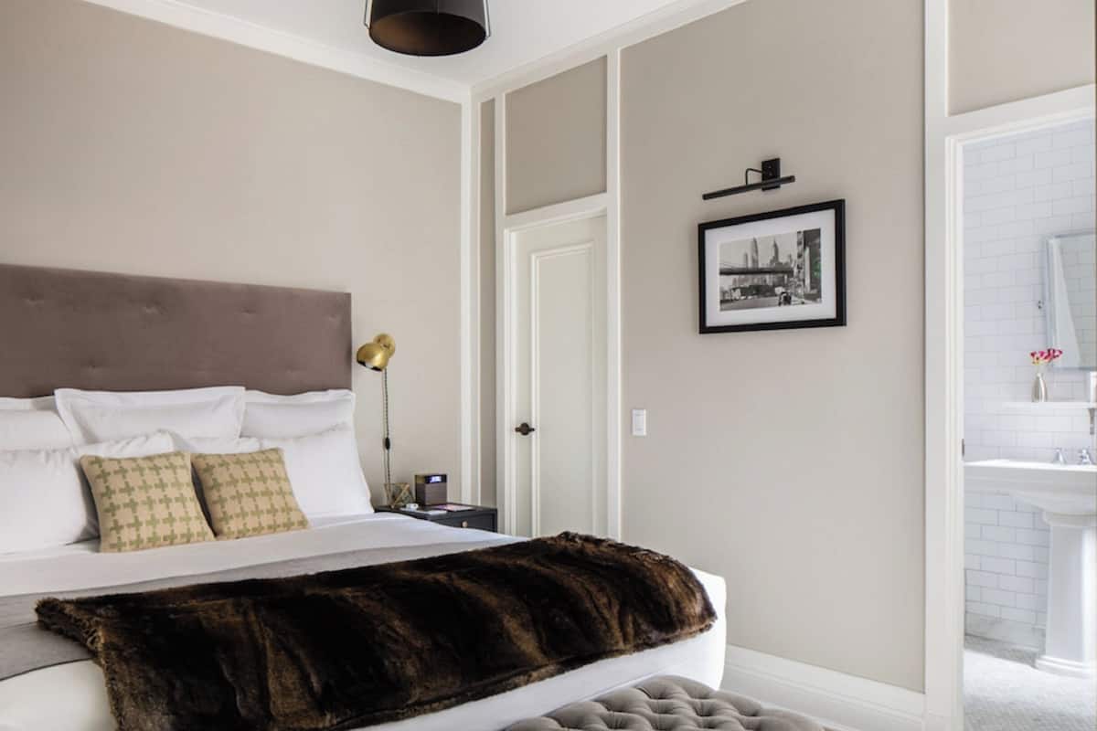 A bedroom with a large bed, a headboard, bedside lamps, a framed picture, and a bathroom visible through an open door.