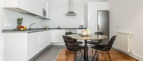 A modern kitchen with white cabinets, a stainless steel refrigerator, a round dining table with four chairs, and a fruit bowl on the counter.