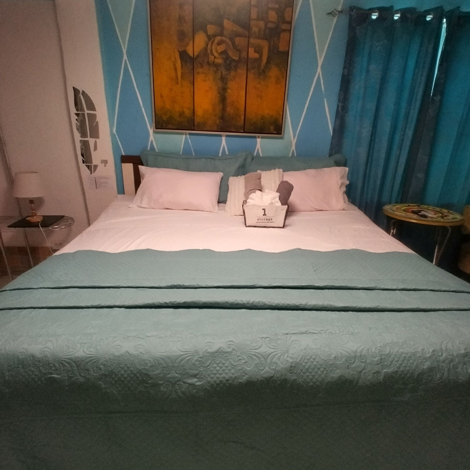 Ein Hotelzimmer mit einem Bett, einem Nachttisch, einer Lampe, einer Wand mit einem Gemälde und einem Vorhang.