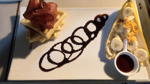 Un plato de postre con una banana, salsa de chocolate y un recipiente pequeño de almíbar.