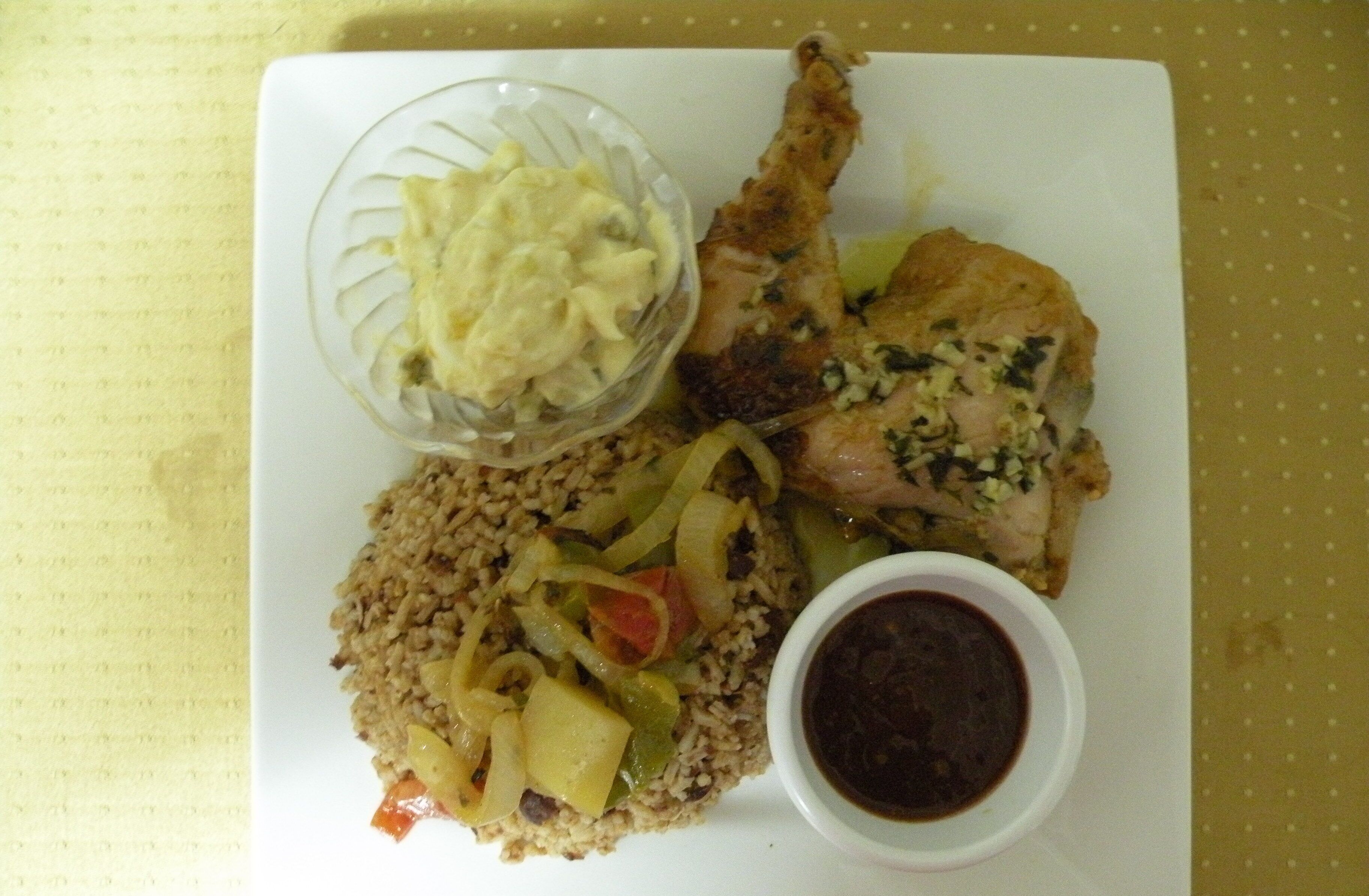 Un plato de comida con arroz, verduras y un trozo de carne, servido sobre un plato blanco con un recipiente pequeño de salsa.