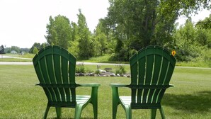 Deux chaises Adirondack vertes sont placées face à une zone herbeuse agrémentée d’arbres et d’un petit élément décoratif aquatique.