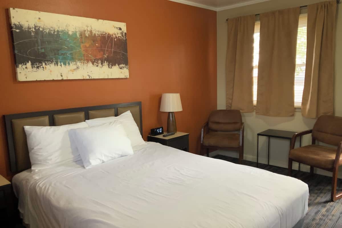 A hotel room with a large bed, a nightstand with a lamp, two chairs, and a painting on the wall.
