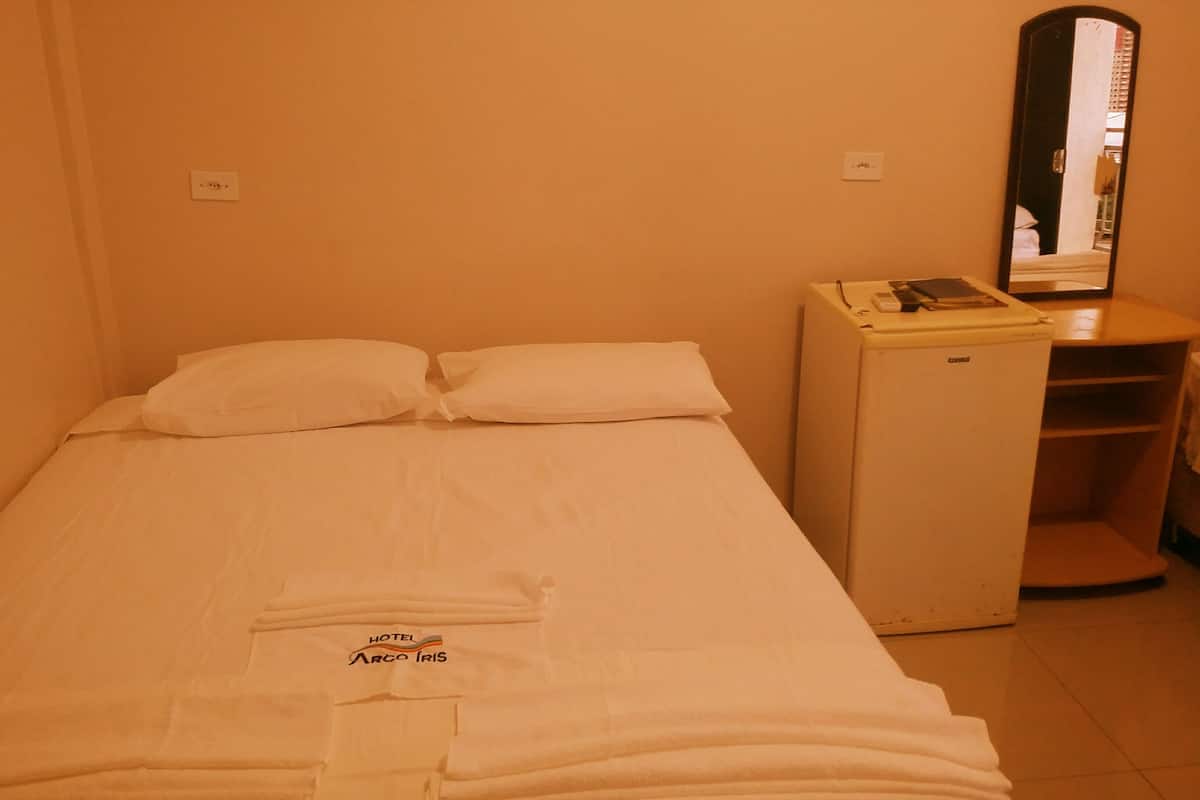 Een hotelkamer met een bed, een klein bureau, een spiegel en een opgerolde handdoek op het bed.