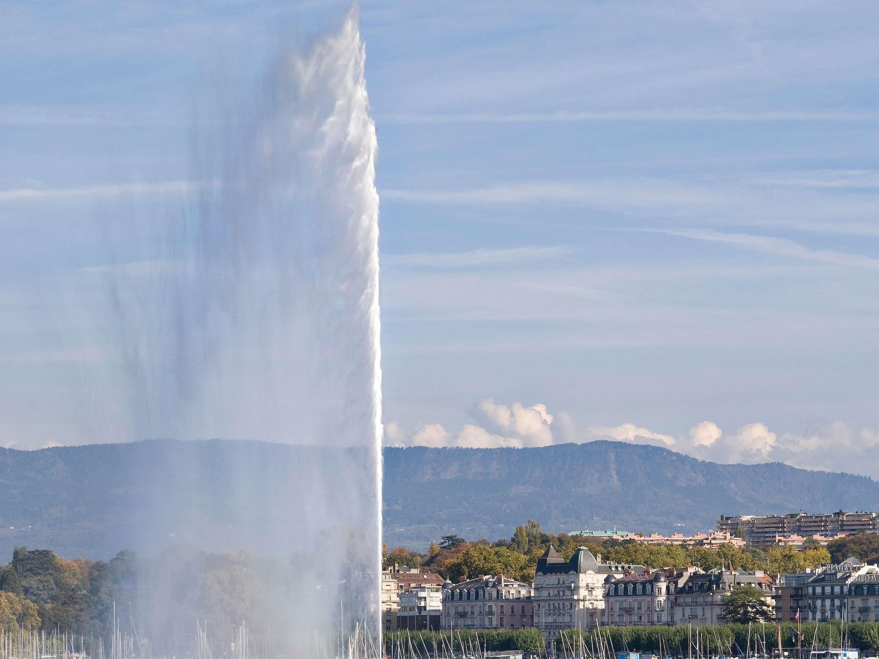 A fountain with water jets in a cityscape with buildings and mountains in the background.