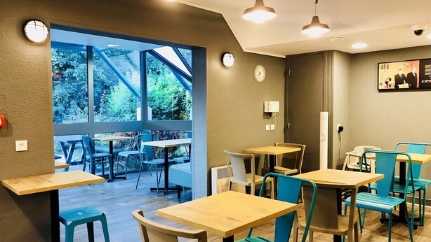 A modern cafe interior with wooden tables and teal chairs, large windows offering a view of greenery, and a clock on the wall. - hotelF1 Geneve Ferney Airport (Ferney-Voltaire)