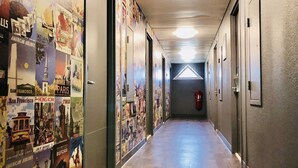 A hallway with posters of cities and landmarks on the walls, a fire extinguisher mounted on the wall, and a ceiling light. - hotelF1 Geneve Ferney Airport (Ferney-Voltaire)