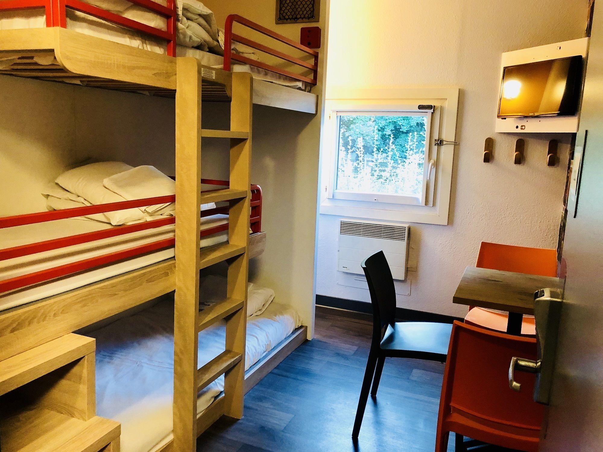 Quarto de dormitório com beliches, uma escrivaninha, uma cadeira, uma janela com vista para árvores e uma TV fixada na parede.