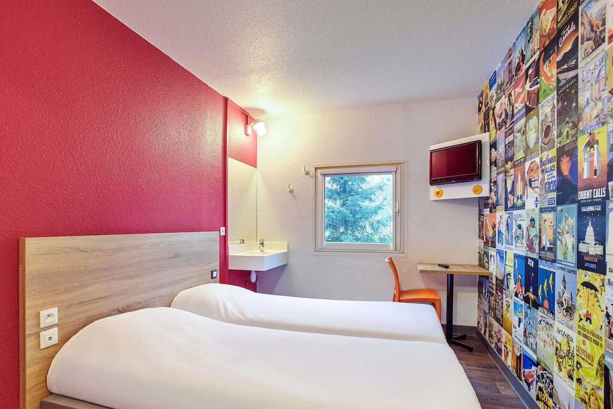 A hotel room with a bed, a desk with a TV, a window, and a wall decorated with travel posters.