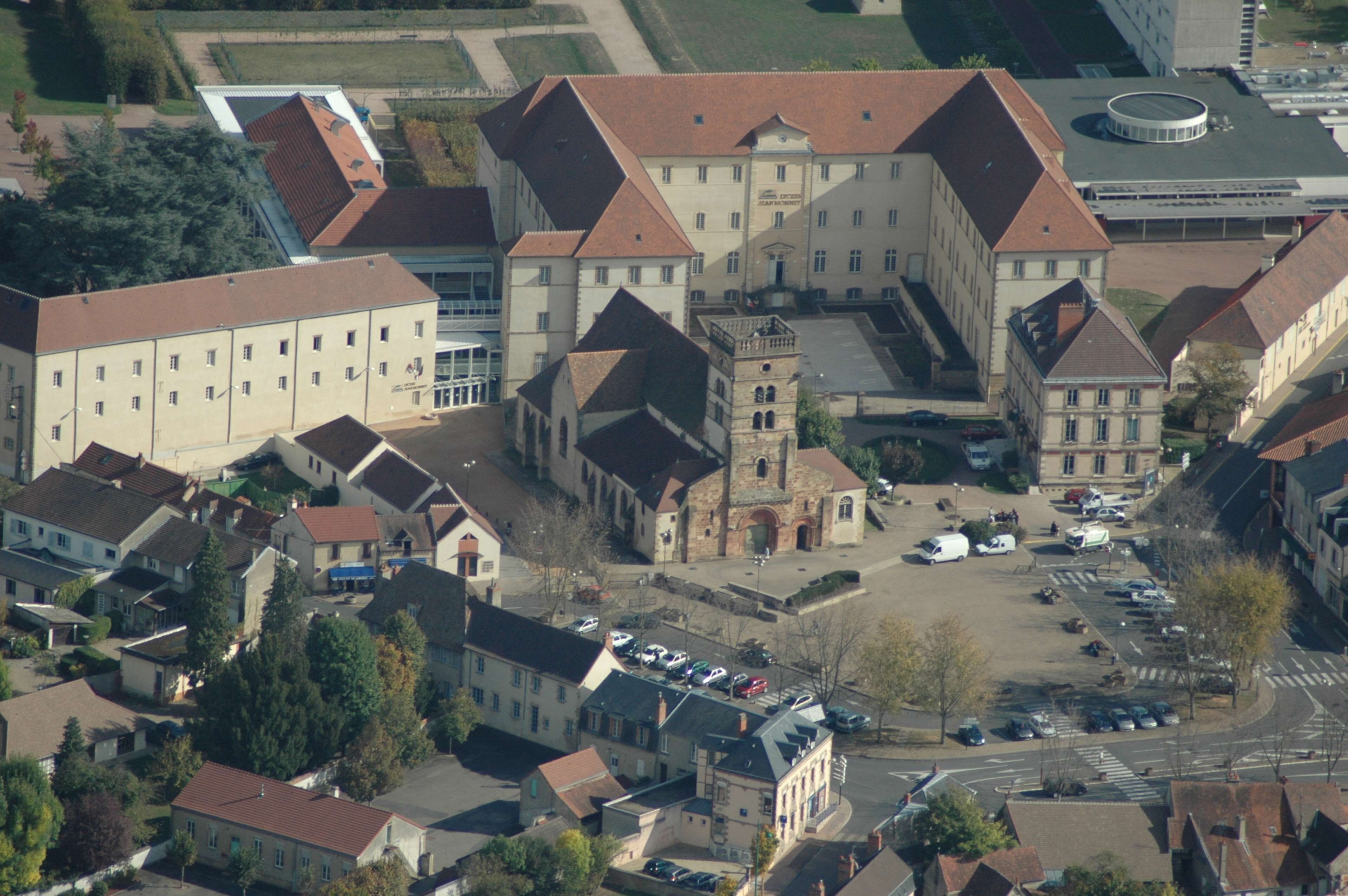 An aerial view of a large building with multiple wings and a central tower, surrounded by smaller buildings and parked cars.