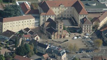 An aerial view of a large building with multiple wings and a central tower, surrounded by smaller buildings and parked cars.