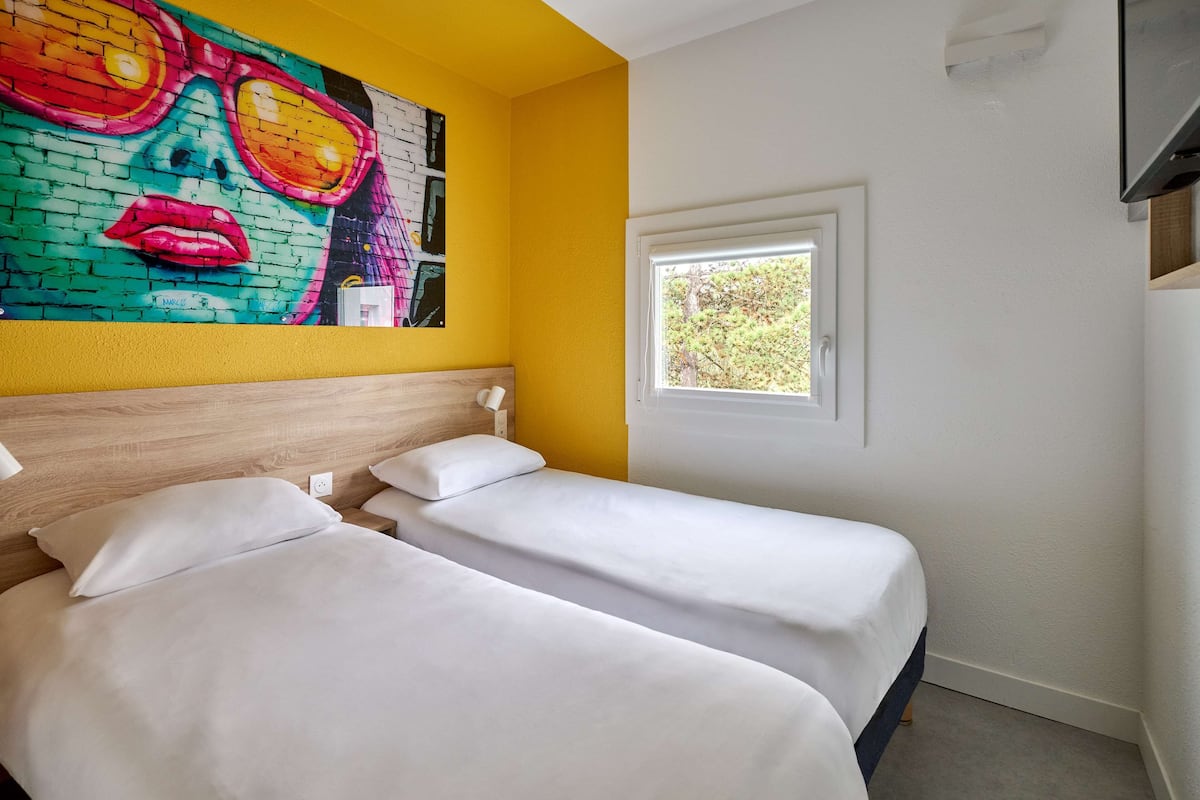 Een hotelkamer met twee bedden, een kleurrijke muurschildering, een raam met uitzicht op bomen en een aan de muur bevestigde televisie.
