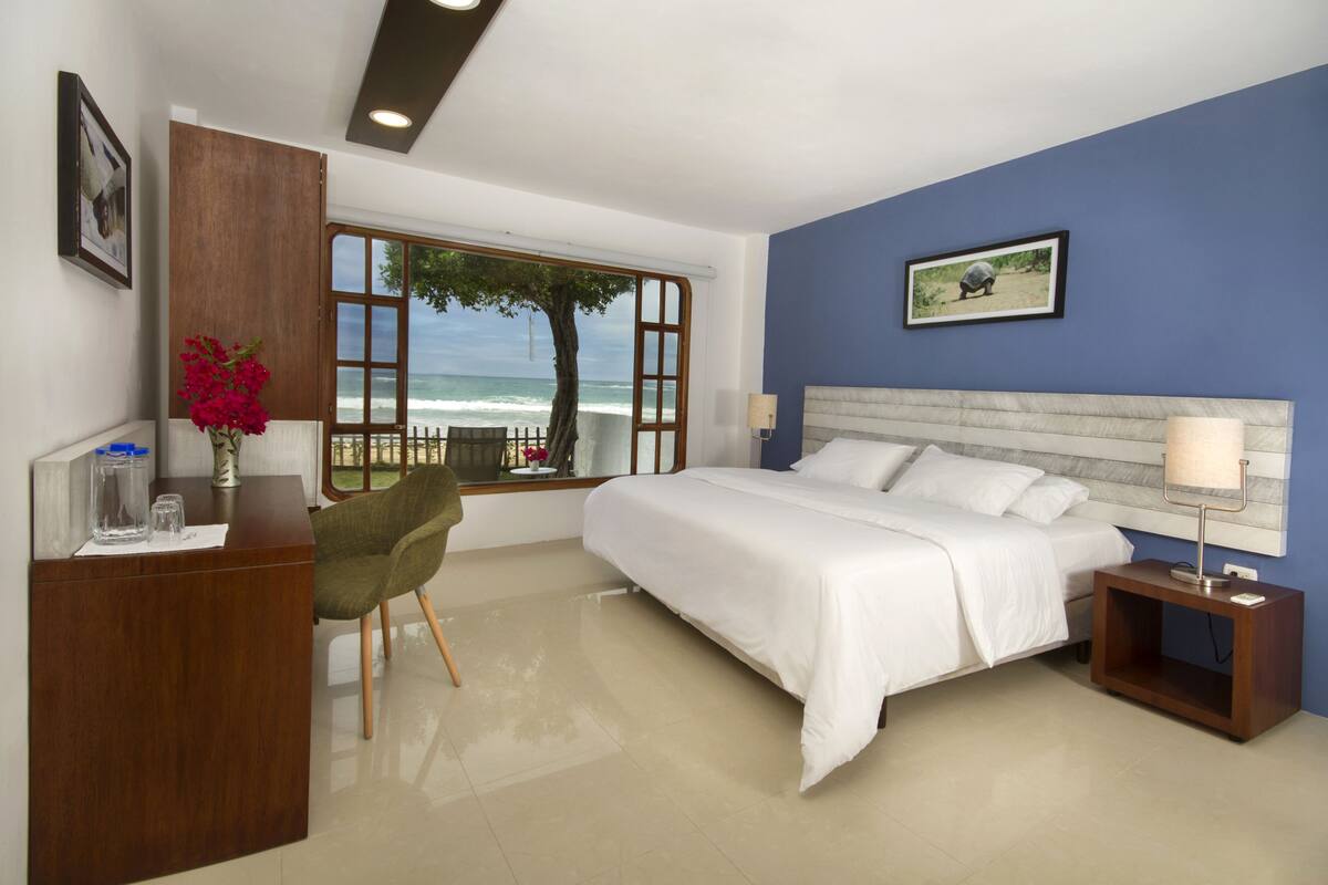 A hotel room with a bed, a desk, a chair, a nightstand, a lamp, a window with a view of the ocean, and a framed picture on the wall.