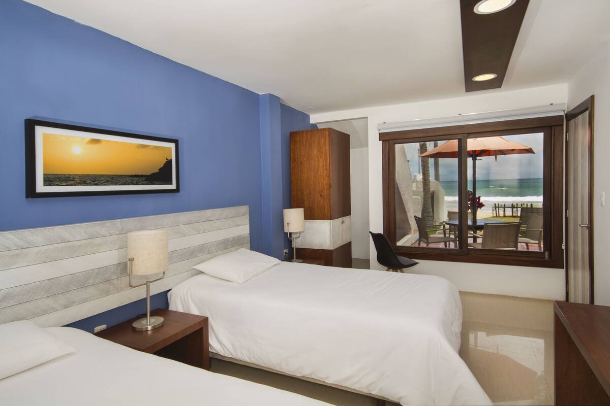 A hotel room with two beds, a view of the ocean through a glass door, a framed sunset photo on the wall, and a wooden cabinet.
