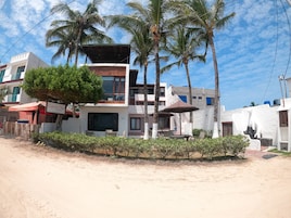 Una propiedad frente a la playa con un edificio de dos pisos, asientos al aire libre, palmeras y una playa de arena.