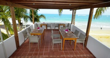 Un espacio al aire libre cubierto con mesas y sillas, con vista a una playa con palmeras y el océano.