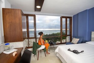 Una habitación con un ventanal grande con vistas a la playa, una cama, un escritorio con una silla y una maleta.